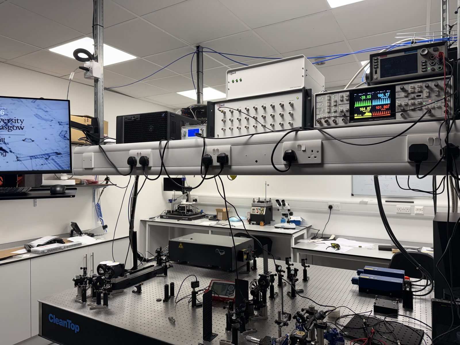 The UltraQUTE laboratory: optical table with the femtosecond laser and optics, measurement electronics on the shelf above, and workbenches behind